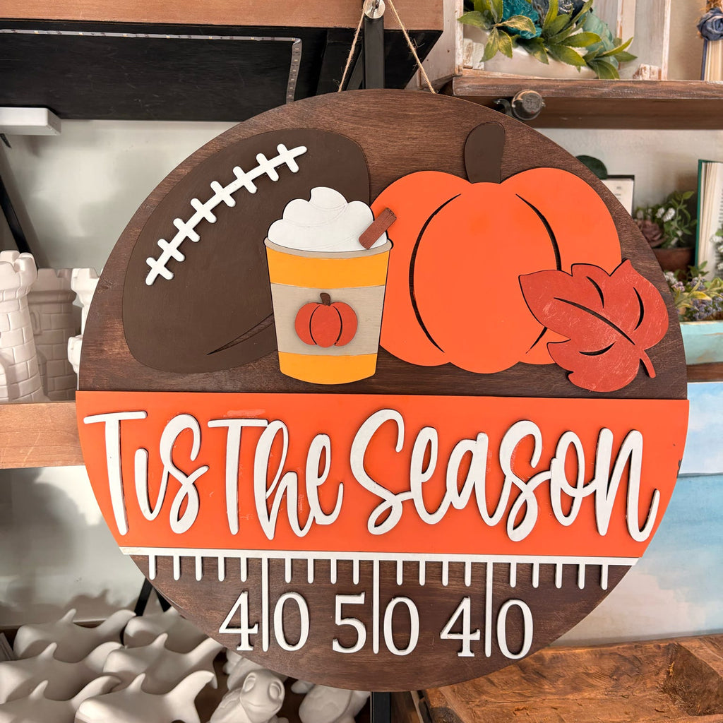A round wooden Tis The Season fall sign hanging on a shelf, featuring a football, pumpkin, hot dog with mustard, autumn leaf, and a measurement ruler at the bottom with the numbers 4, 10, 5, 10, 4, 10.