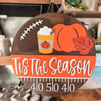 A round wooden Tis The Season fall sign hanging on a shelf, featuring a football, pumpkin, hot dog with mustard, autumn leaf, and a measurement ruler at the bottom with the numbers 4, 10, 5, 10, 4, 10.