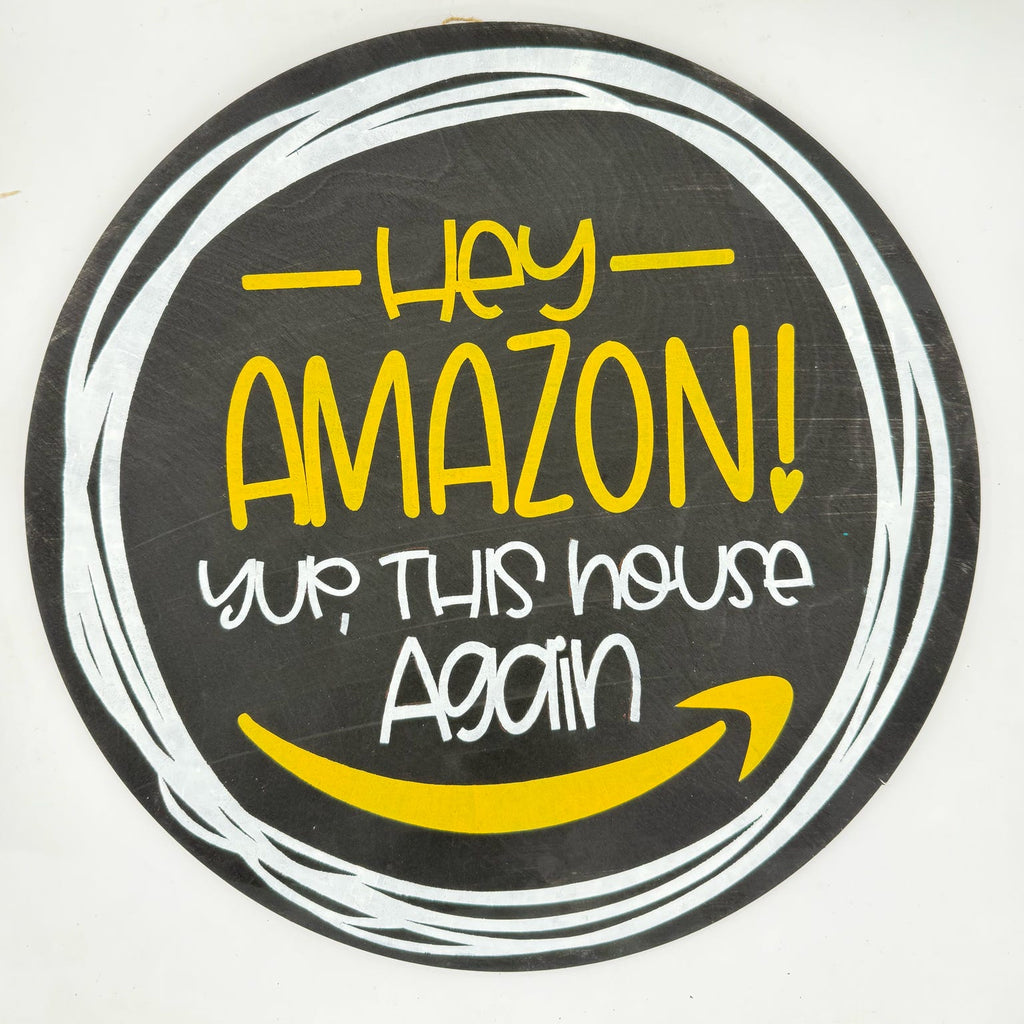 A circular novelty sign featuring a black background with white hand-drawn circle lines, yellow text reading 'Hey AMAZON! yup, THIS house Again', and a yellow smiley face inspired by the Amazon logo.