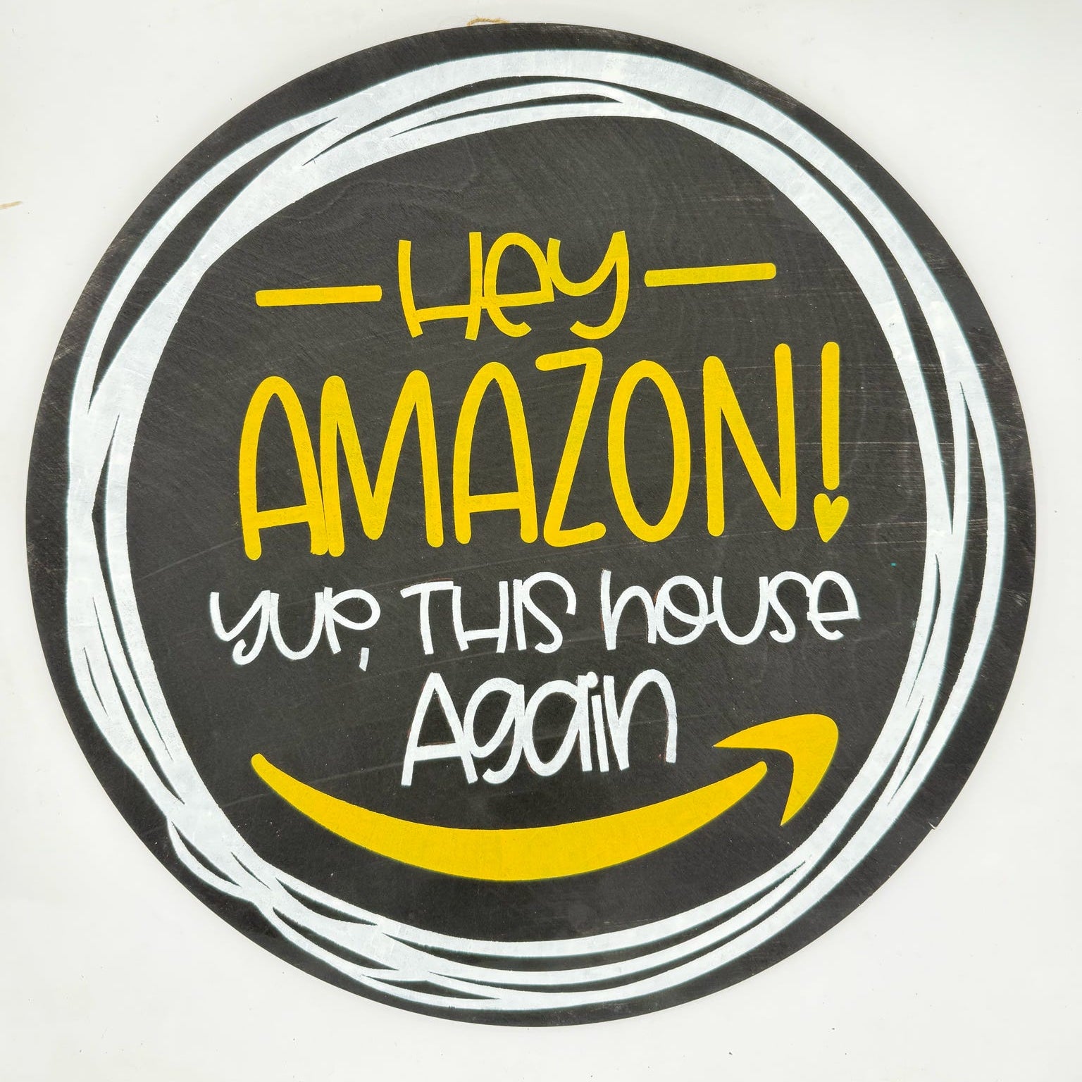 A circular novelty sign featuring a black background with white hand-drawn circle lines, yellow text reading 'Hey AMAZON! yup, THIS house Again', and a yellow smiley face inspired by the Amazon logo.