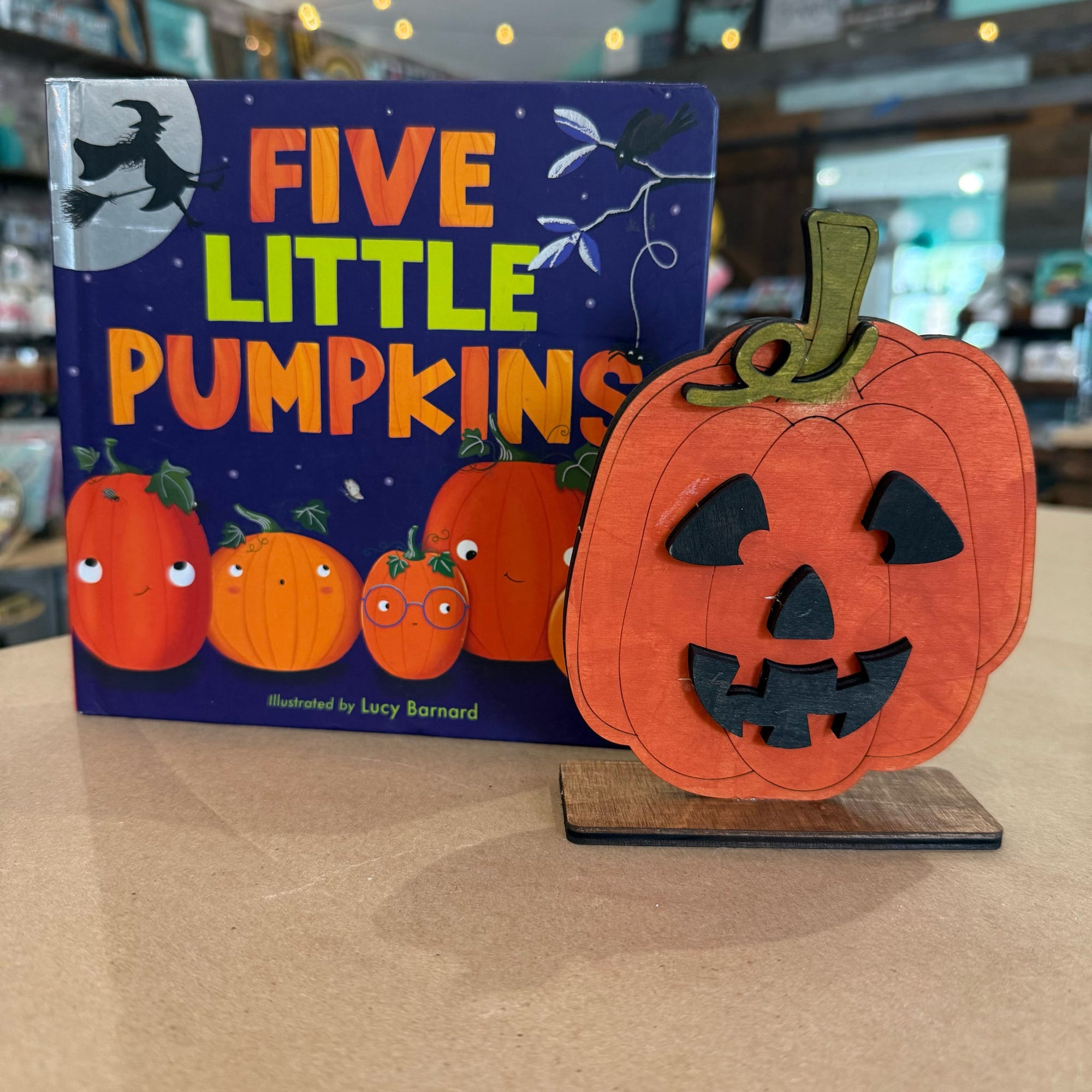 A children's book titled "Five Little Pumpkins" by Lucy Barnard stands next to a carved wooden jack-o'-lantern decoration on a tabletop in a store setting.