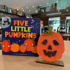 A children's book titled "Five Little Pumpkins" by Lucy Barnard stands next to a carved orange pumpkin decoration on a wooden stand in a softly lit indoor room.