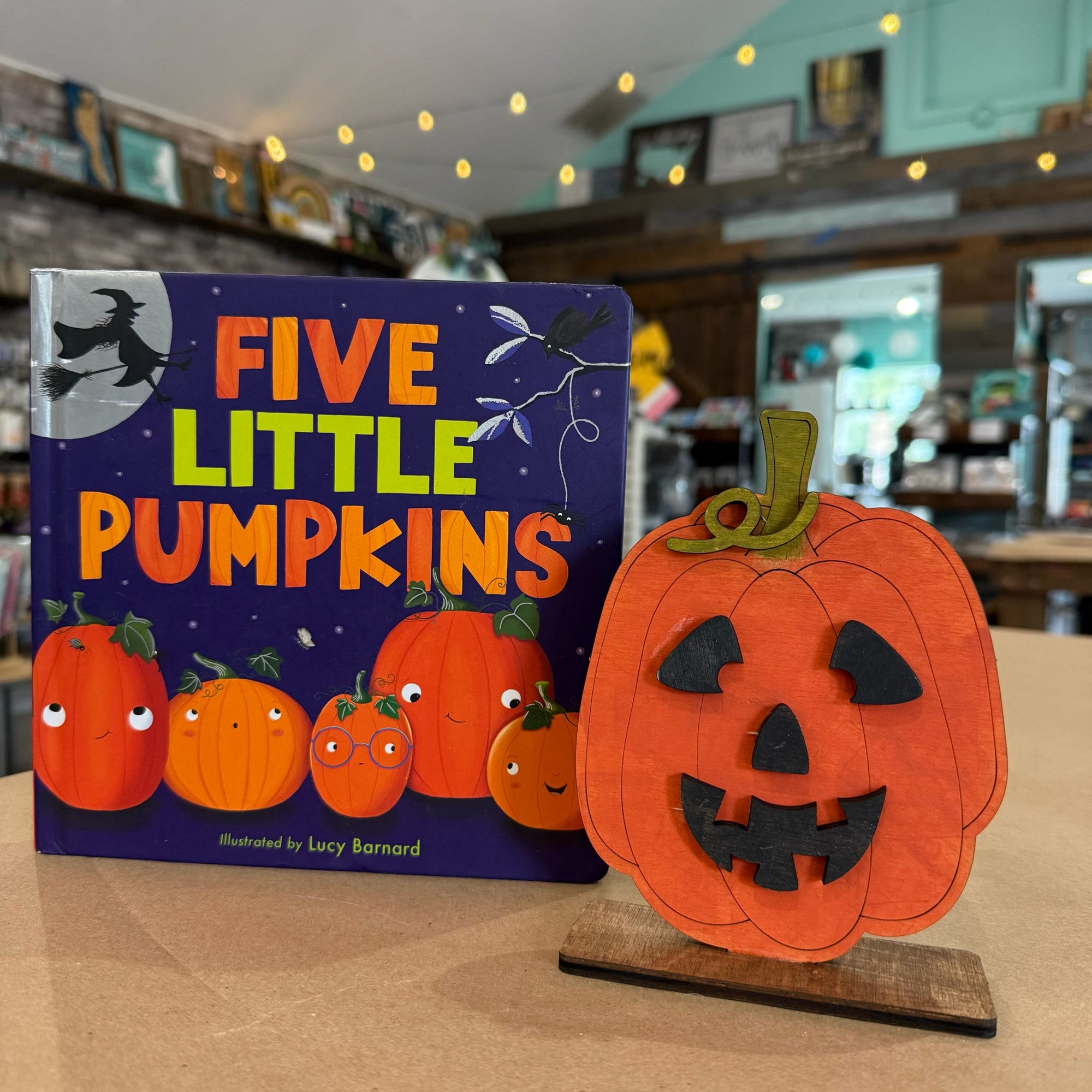 A children's book titled "Five Little Pumpkins" by Lucy Barnard stands next to a carved orange pumpkin decoration on a wooden stand in a softly lit indoor room.