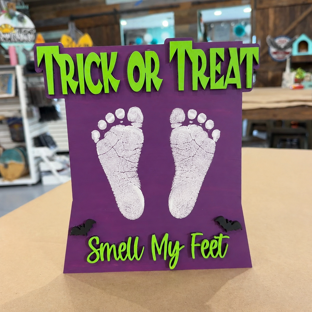 A Halloween novelty card or sign with a purple background, bright green 'TRICK OR TREAT' text at the top, white baby footprints in the center, small black bat shapes, and green 'Smell My Feet' text at the bottom, displayed on a tabletop with a blurred store background.