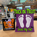 Two Halloween treats displayed on a counter in a shop: a square black box with orange and purple text reading 'DON'T PUSH THE BUTTON! A Halloween Treat' by Smell Like Coffee, and a purple card with white footprints and green text reading 'TRICK OR TREAT' and 'Smell My Feet'.