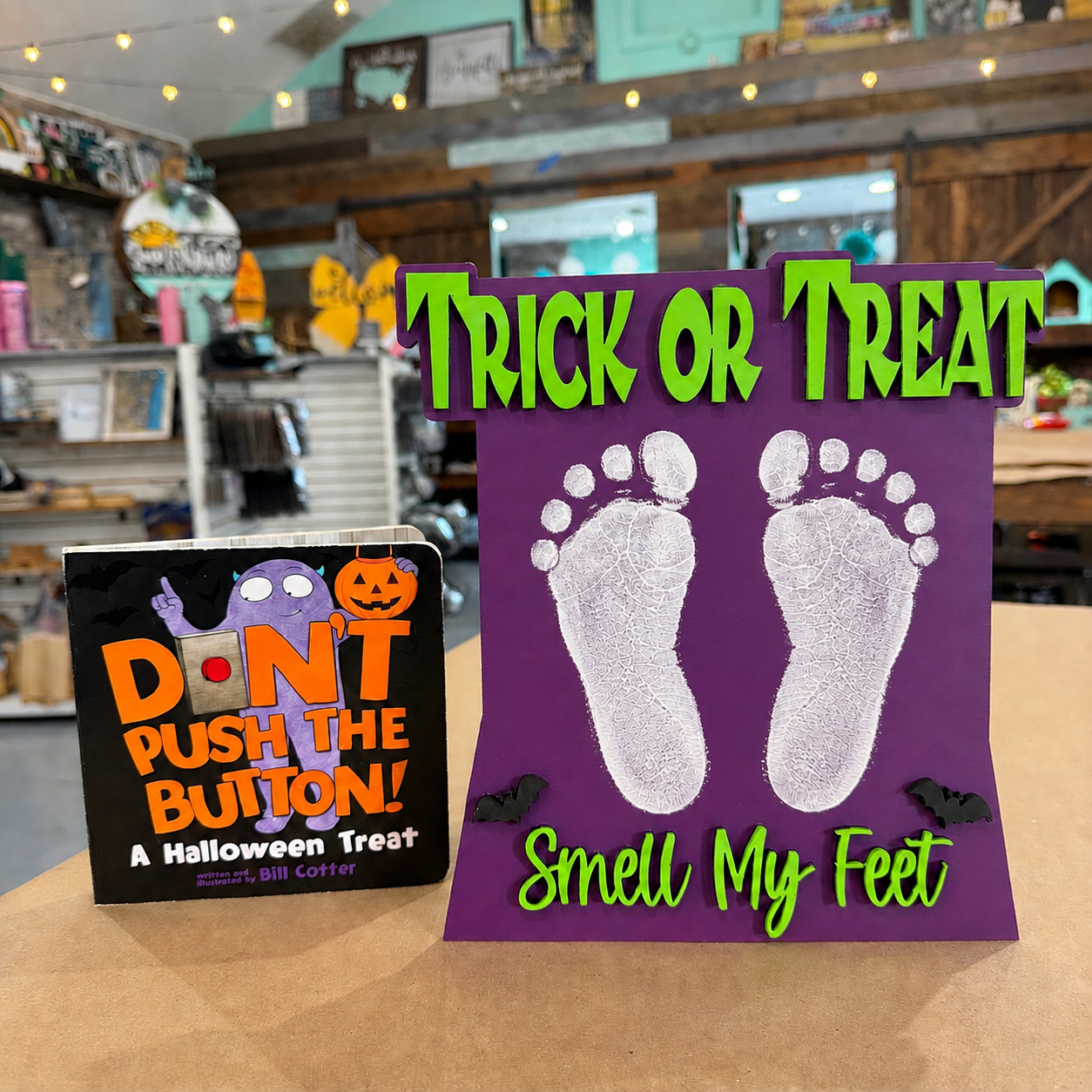 Two Halloween treats displayed on a counter in a shop: a square black box with orange and purple text reading 'DON'T PUSH THE BUTTON! A Halloween Treat' by Smell Like Coffee, and a purple card with white footprints and green text reading 'TRICK OR TREAT' and 'Smell My Feet'.