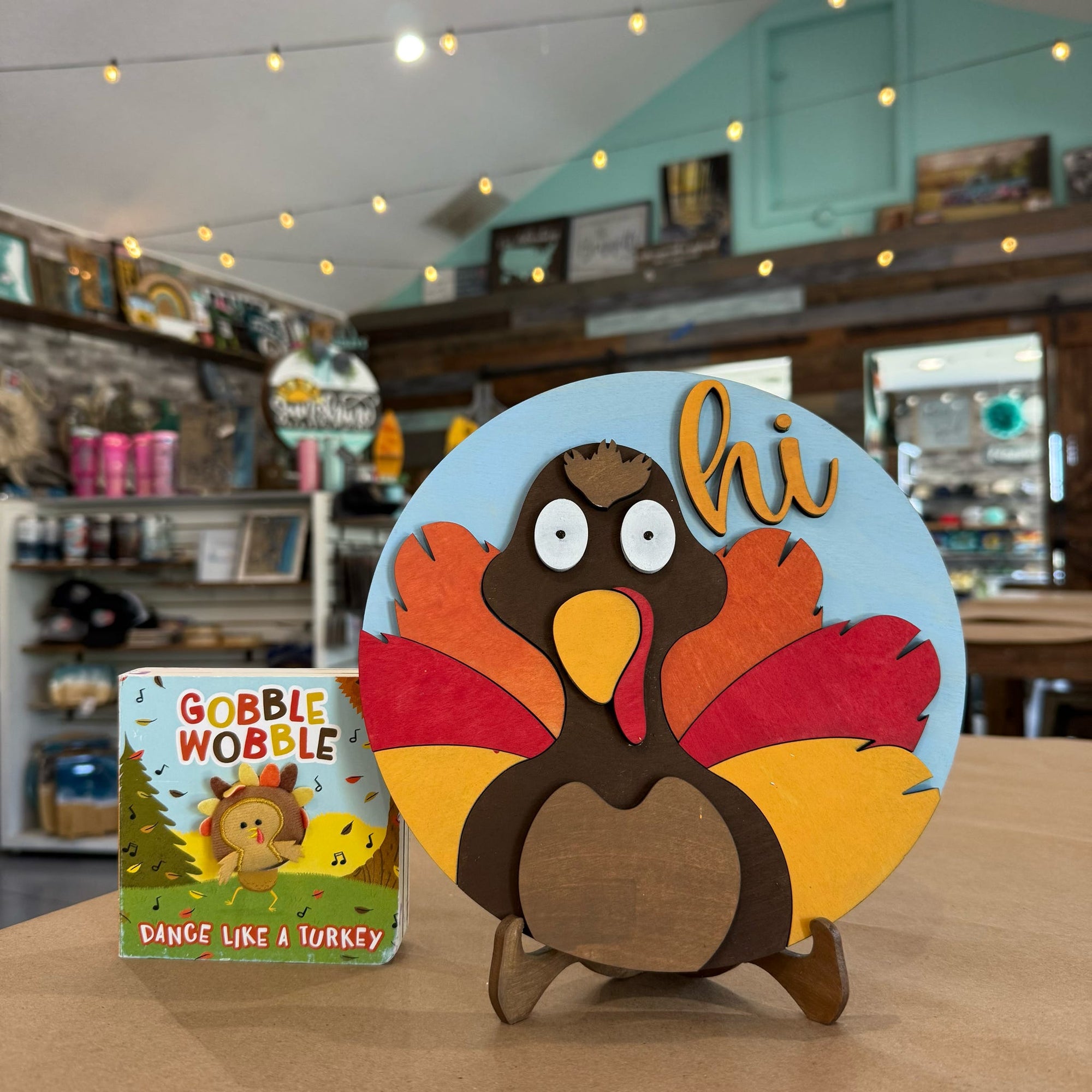 A colorful wooden turkey greeting sign on a small stand, displayed on a table inside a shop. The sign features a brown turkey with red, orange, and yellow feathers on a light blue circular background with the word 'hi' in orange.