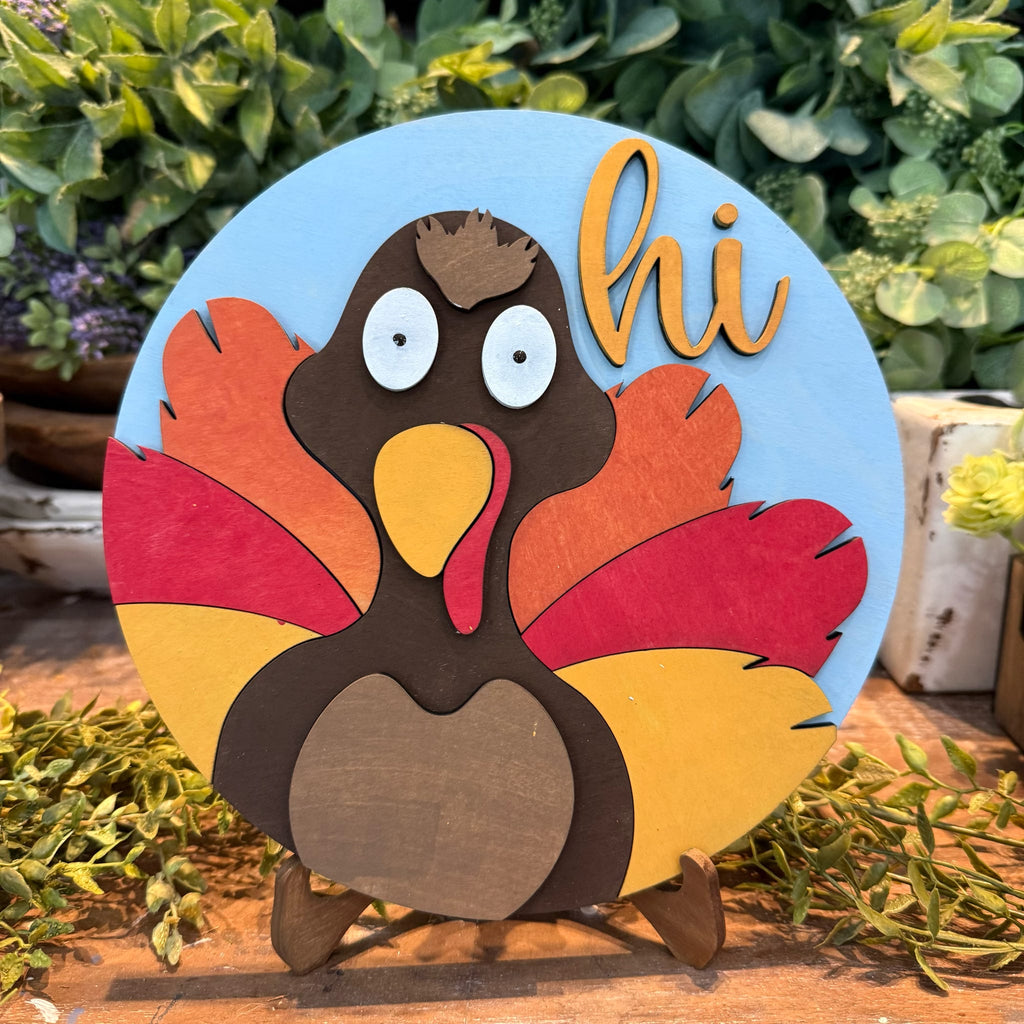 A circular wooden Thanksgiving decoration featuring a stylized turkey with layered brown, red, orange, and yellow feathers on a light blue background, with the word "hi" in orange script. The sign is displayed on a small wooden stand outdoors among green plants.