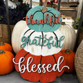 A door hanger with three pumpkin shapes and accompanying text that reads 'THANKSGIVING', 'GRATEFUL', and 'BLESSED', displayed in a store with pumpkins in the background.