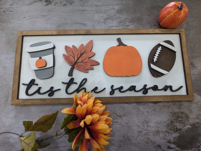 A 3D sign with a football, pumpkin, and leaves design, with the phrase 'tis the season' written on it, displayed on a table with autumn decorations around it.