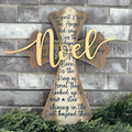 A wooden cross with gold paint, featuring an angel's kiss and the words 'Noel' in the center, mounted on a brick wall with greenery at the bottom.