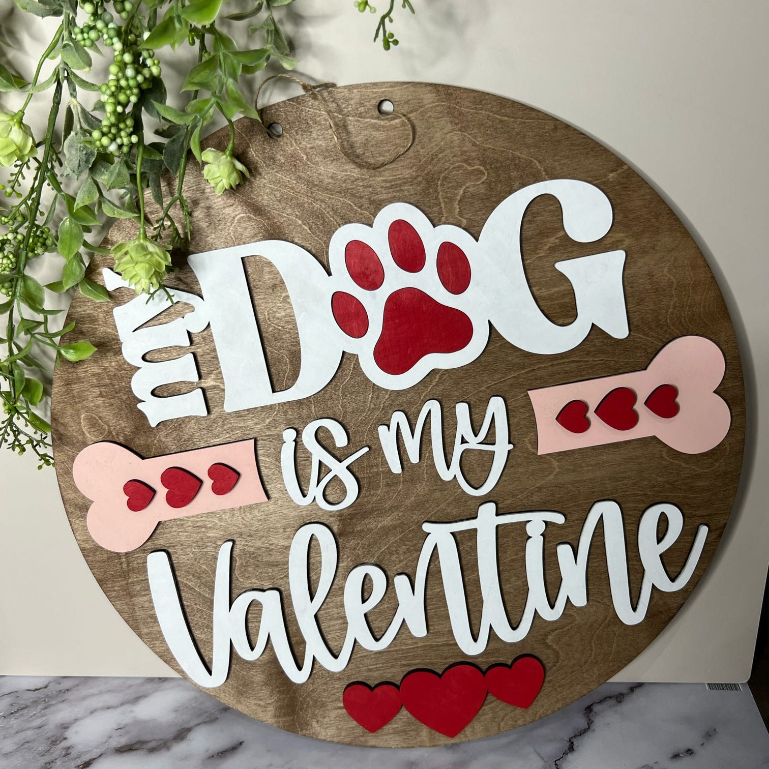 A wooden door hanger with the phrase 'MY DOG IS MY VALENTINE' written in whimsical lettering, accompanied by illustrations of a dog paw and bones. The background is a neutral color, and there are decorative elements in the top right corner.