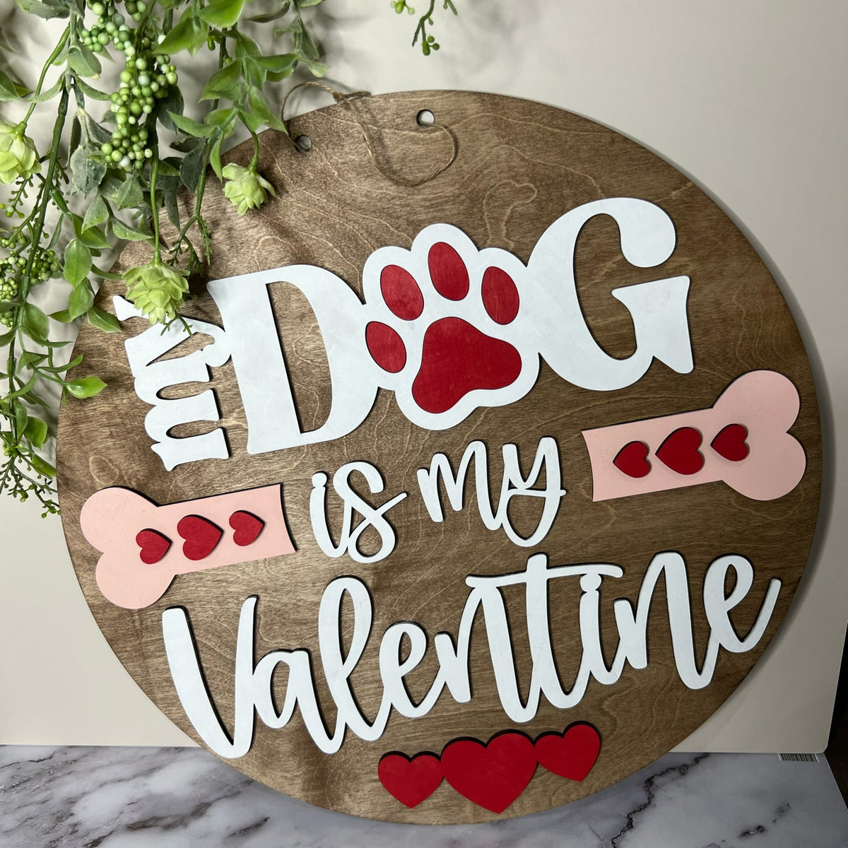 A wooden door hanger with the phrase 'MY DOG IS MY VALENTINE' written in whimsical lettering, accompanied by illustrations of a dog paw and bones. The background is a neutral color, and there are decorative elements in the top right corner.