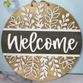 A circular wooden door hanger with a cut-out word 'WELCOME' in the center, decorated with herb shoot designs around the border.