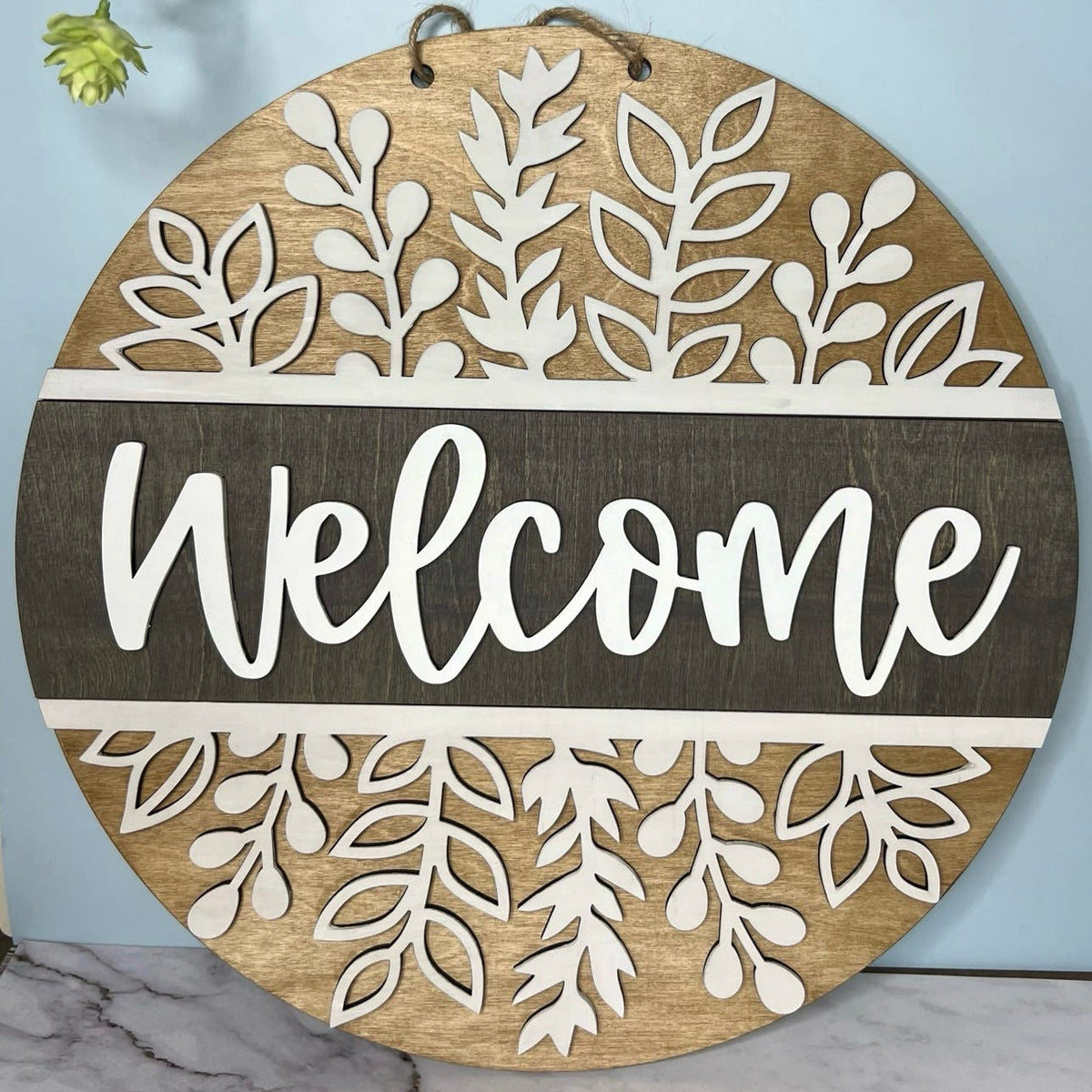 A circular wooden door hanger with a cut-out word 'WELCOME' in the center, decorated with herb shoot designs around the border.
