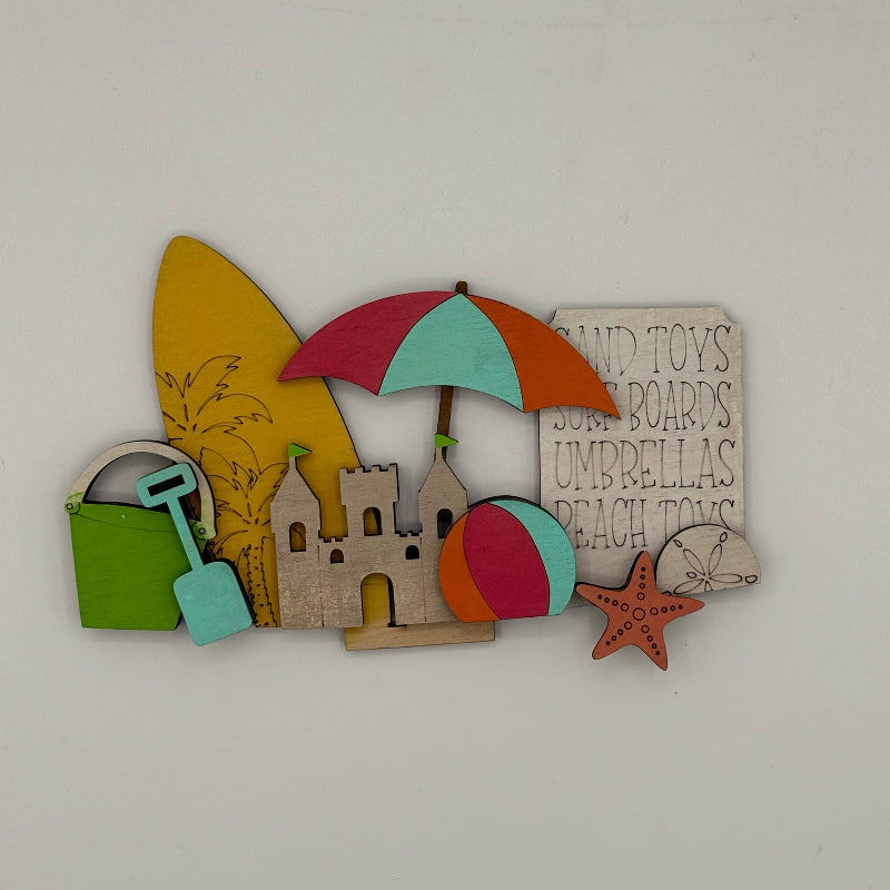 Decorative wall art with beach-themed items including an umbrella, sandcastle, and surfboard on a plain background.