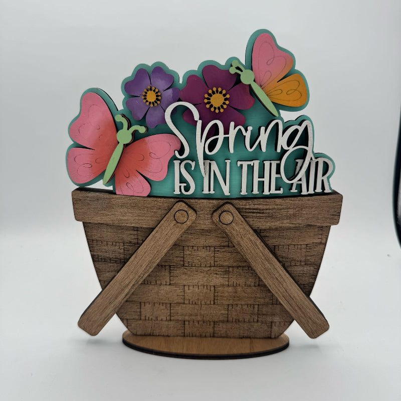 Decorative sign with 'Spring is in the Air' text, butterflies, and flowers on a wooden basket stand.