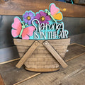 Decorative sign with 'Spring is in the Air' text, flowers, and butterflies on a wooden surface.