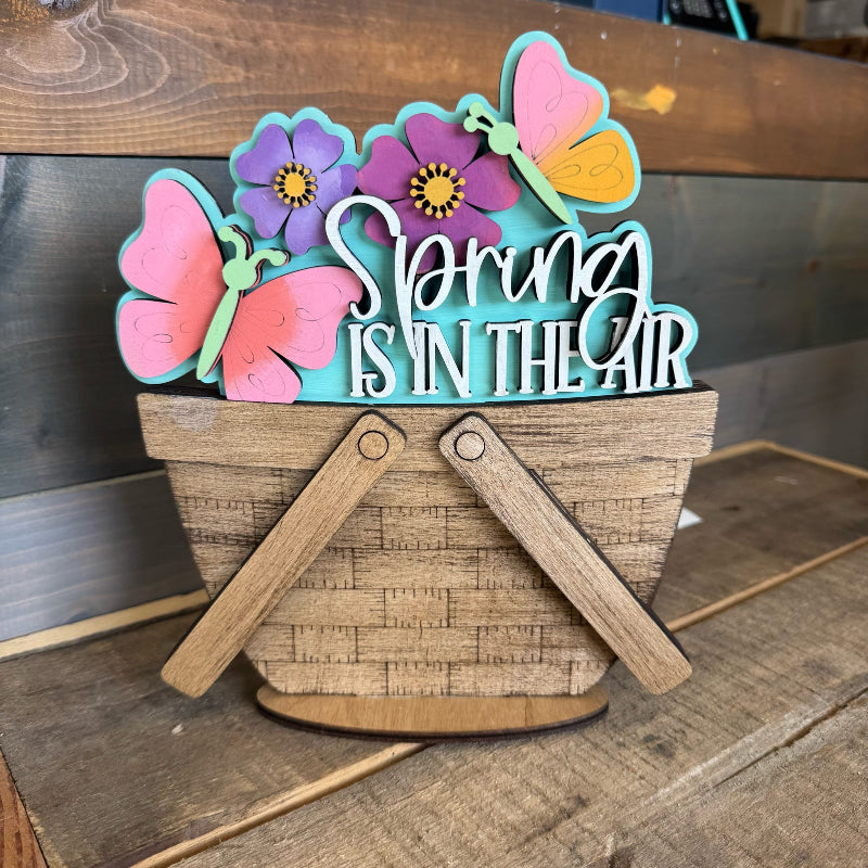 Decorative sign with 'Spring is in the Air' text, flowers, and butterflies on a wooden surface.
