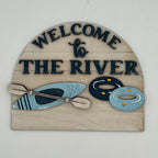 Decorative sign with 'Welcome to The River' text and kayak, paddle, and life jacket illustrations on a white background.