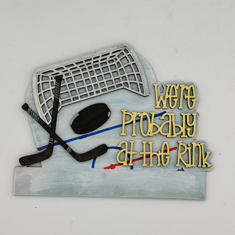 Decorative sign with hockey equipment and text 'We're Probably at the Rink' on a white background