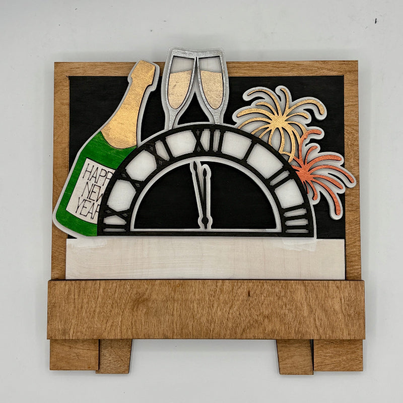 Decorative New Year's Eve sign with champagne bottle, glasses, clock, and fireworks on a wooden base.