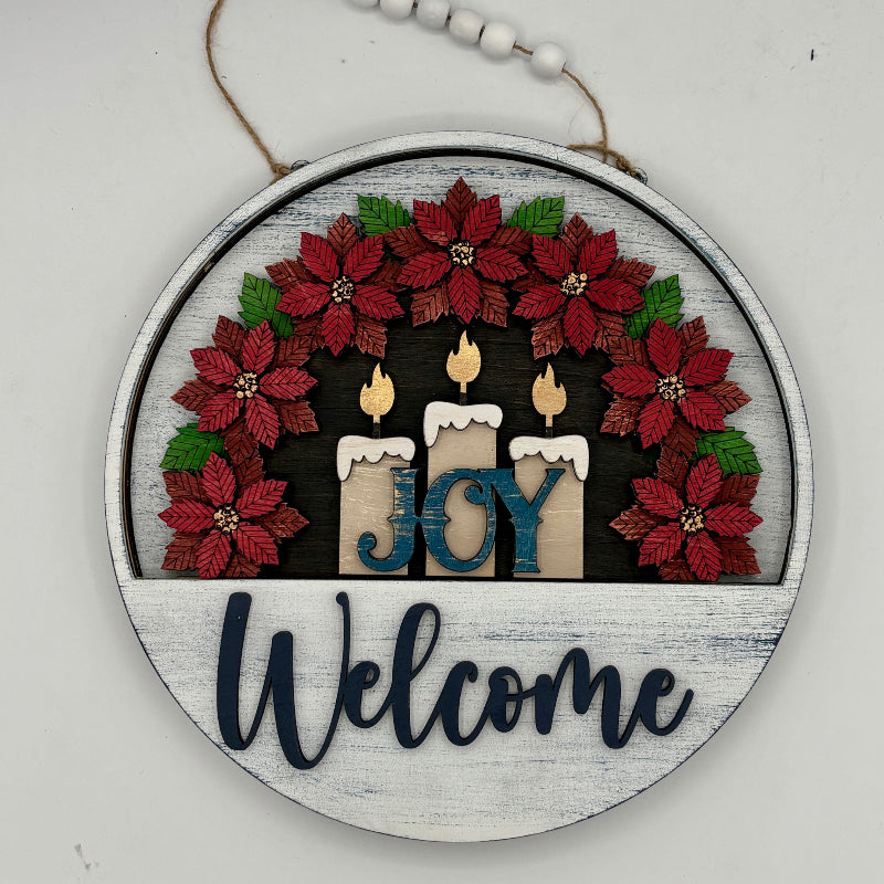 Decorative sign with candles and poinsettias, featuring the word 'JOY' and the word 'Welcome'.