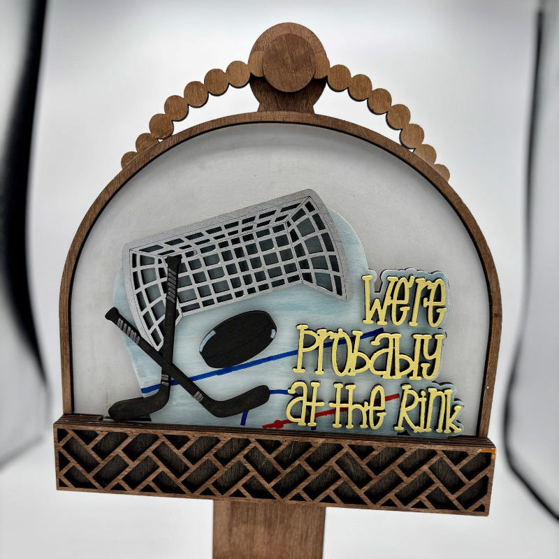 Decorative sign with hockey theme and text 'We're Probably at the Rink' on a stand.