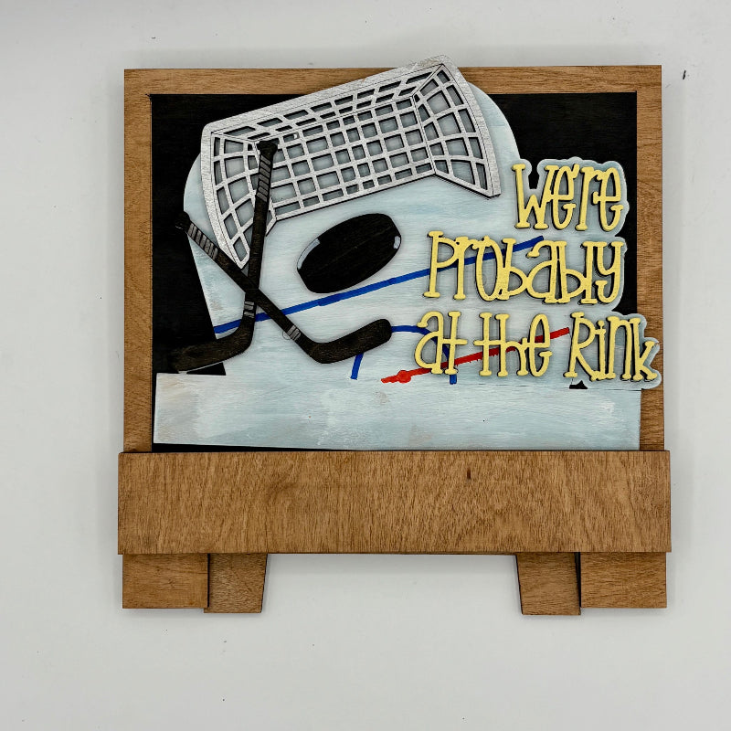 Decorative sign with hockey elements and text 'We're Probably at the Rink' on a white background