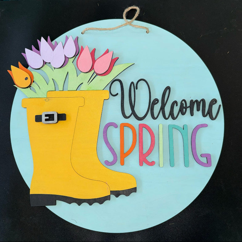 Decorative sign with a yellow rain boot, flowers, and 'Welcome Spring' text on a black background.