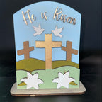 Decorative item with crosses, lilies, doves, and 'He is Risen' text on a blue background.