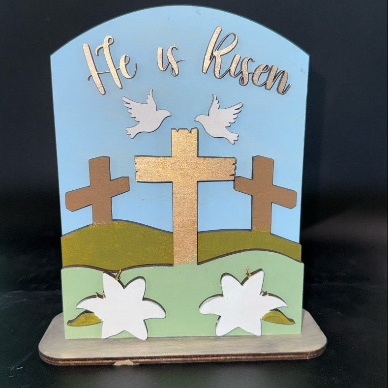 Decorative item with crosses, lilies, doves, and 'He is Risen' text on a blue background.