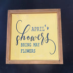 Wooden-framed sign with 'April Showers Bring May Flowers' text on a yellow background