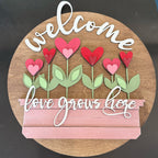 Decorative sign with 'welcome' and 'love grows here' text, featuring heart-shaped flowers on a wooden background.
