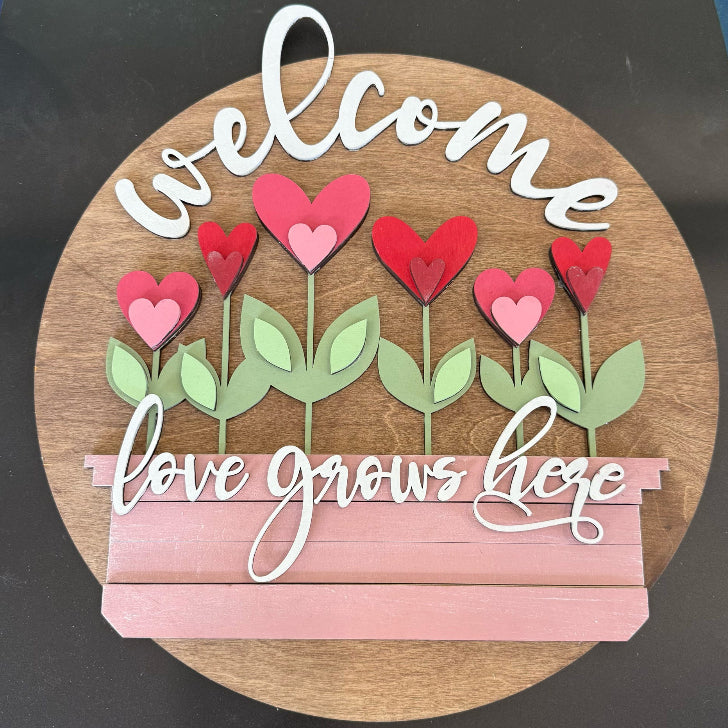 Decorative sign with 'welcome' and 'love grows here' text, featuring heart-shaped flowers on a wooden background.