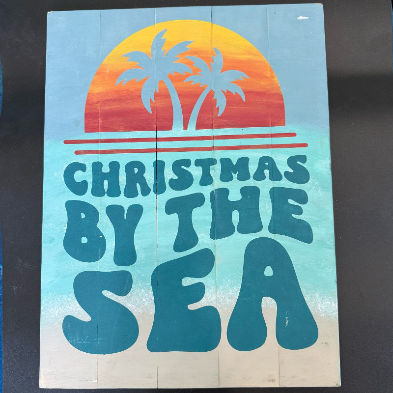 Wooden sign with 'Christmas by the Sea' text and palm tree design on a gradient background.
