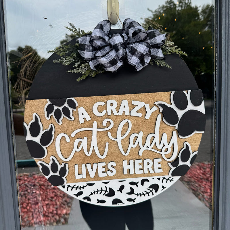 Decorative door hanger with 'A Crazy Cat Lady Lives Here' text and paw prints, attached to a glass door.