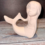 Unfinished ceramic mermaid figurine on a wooden surface with a dark background