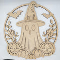 A light natural wood Halloween decorative cutout featuring a ghost in a witch hat, surrounded by bats, flowers, pumpkins, and a circular frame, photographed against a plain light background.