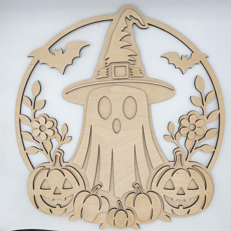 A light natural wood Halloween decorative cutout featuring a ghost in a witch hat, surrounded by bats, flowers, pumpkins, and a circular frame, photographed against a plain light background.