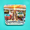 A square coaster with rounded corners featuring a colorful watercolor illustration of Fernandina Beach, Florida, with the text 'Fernandina Beach FLORIDA' at the bottom, shown on a solid turquoise background.