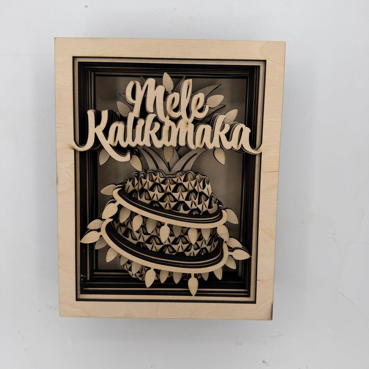 A square wooden wall art piece featuring a laser-cut 3D papercalix design of a kaliko fish with the words 'Mele Kaliko' in raised script above it, mounted on a light wood frame against a plain white wall.