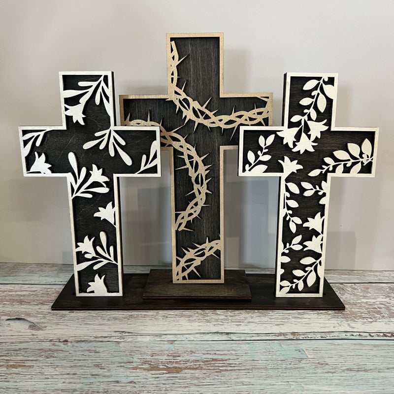 A layered cross decoration featuring three crosses arranged vertically with a floral and barbed wire design, predominantly in black and white, placed on a wooden surface.