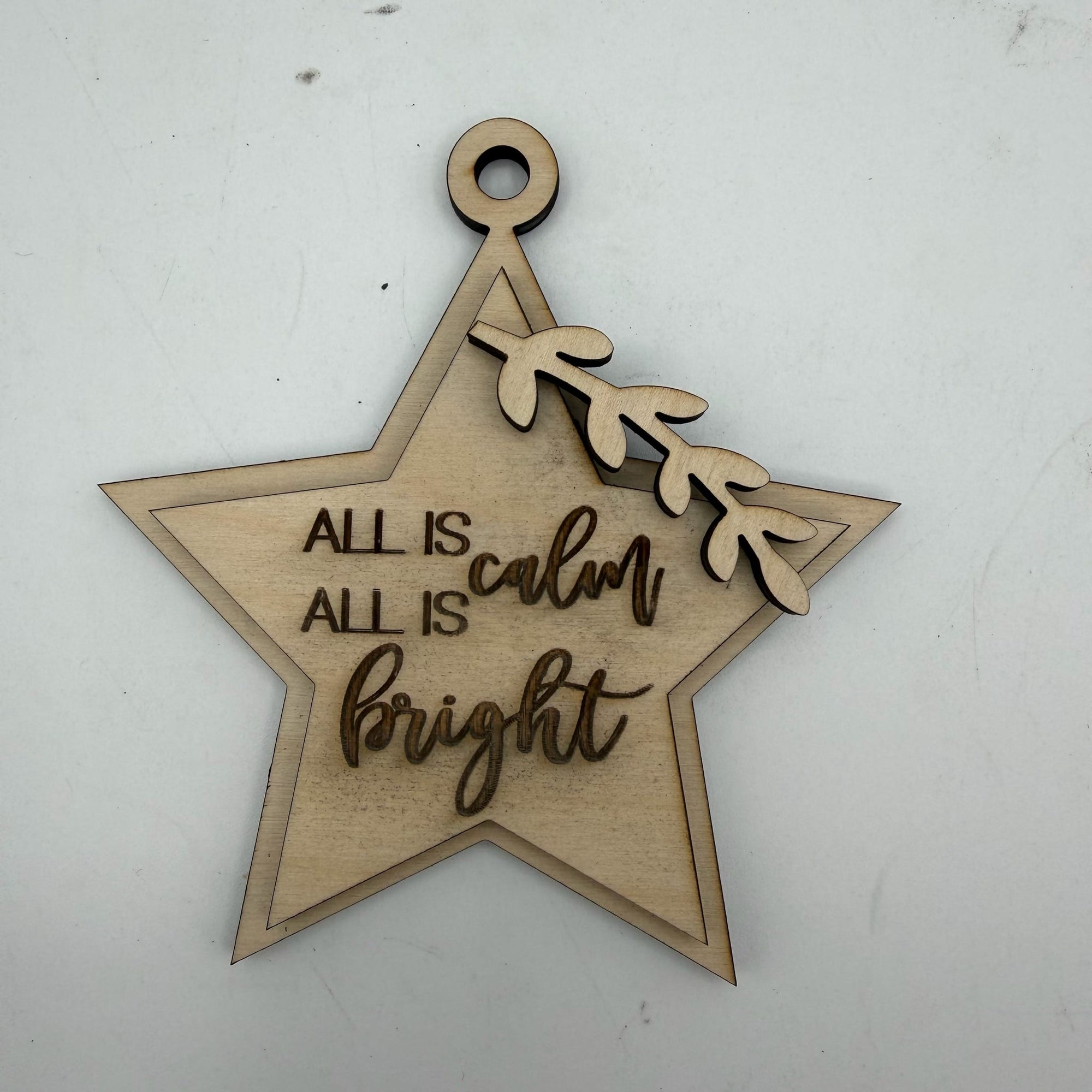 A laser-cut wooden star ornament with the phrase 'ALL IS calm bright' in a mix of block and cursive fonts, decorated with a small leafy branch detail, shown on a light gray background.