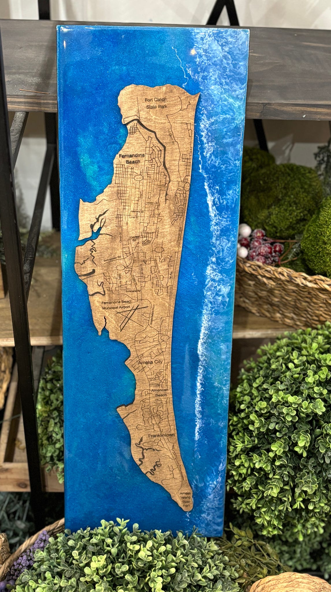 A vertical rectangular wooden plaque featuring a raised map design of Israel, with a natural wood-toned landmass and a bright blue resin-encased background, displayed in a decorative setting with greenery and shelving.