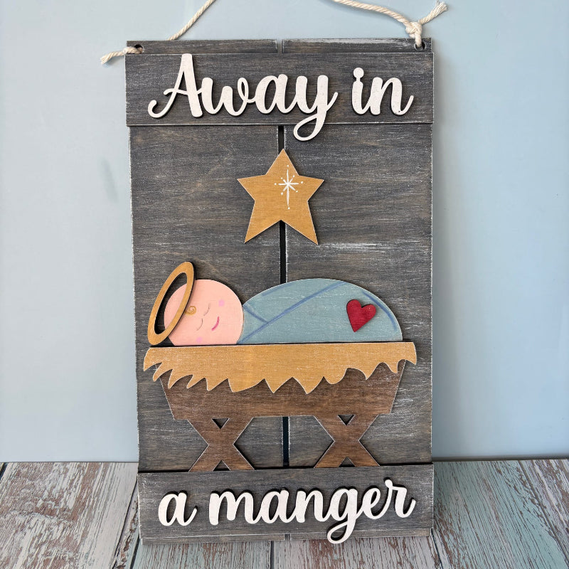 Decorative sign with baby Jesus in a manger and text 'Away in a manger' on a wooden background.