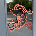 An orange shrimp-shaped door hanger made of wood, displayed on a glass door with a tree visible in the background.
