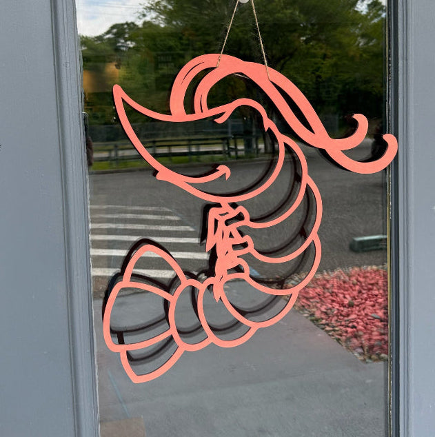 An orange shrimp-shaped door hanger made of wood, displayed on a glass door with a tree visible in the background.