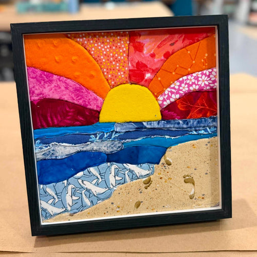 A framed square art piece featuring a colorful quilted sunset beach scene with orange and red sky, yellow sun, blue ocean, and sandy beige shore, displayed on a cardboard surface.
