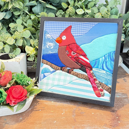 A framed handmade quilted art piece featuring a red cardinal perched on a branch against a blue and light blue quilted background, displayed on a wooden surface with green foliage and red roses in the background.